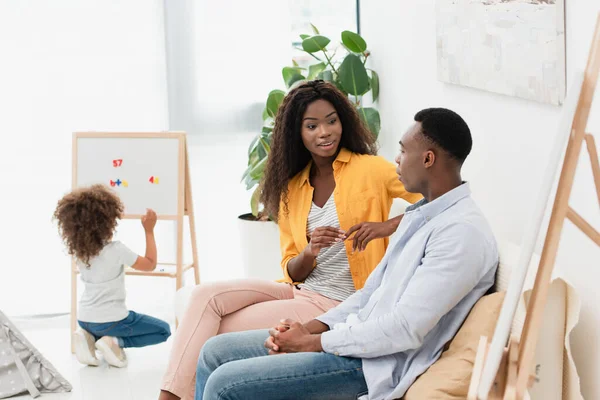 Enfoque selectivo de los padres afroamericanos sentados en el sofá, mientras que la hija rizada sentado cerca del caballete magnético - foto de stock