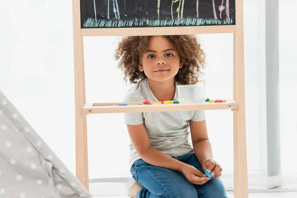 Curly african american kid looking at camera through wooden frame and holding magnet — Stock Photo