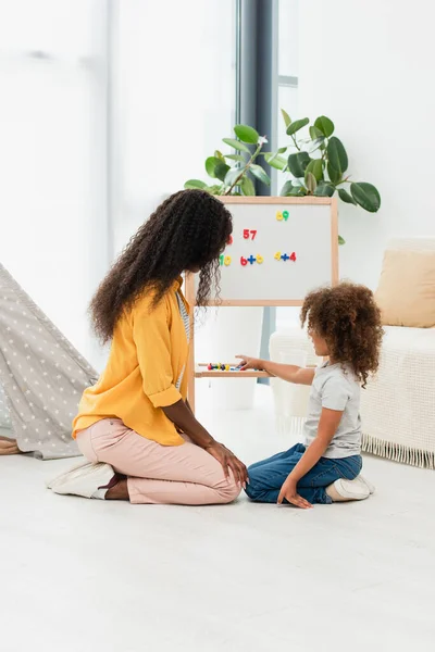 Afro americano figlia punta con dito mentre seduto vicino lavagna e madre — Foto stock