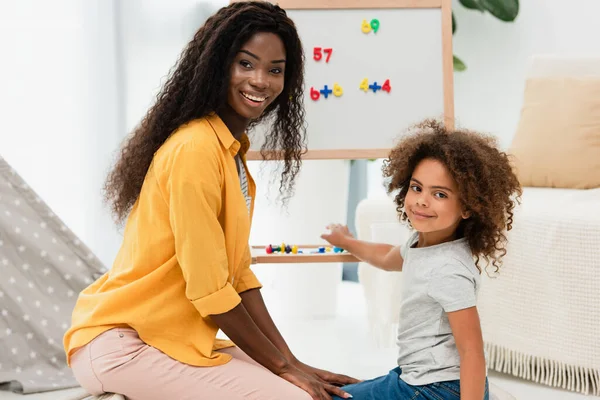 Lockige afrikanisch-amerikanische Mutter und Tochter, die in der Nähe von Whiteboard mit Magneten in die Kamera schauen — Stockfoto