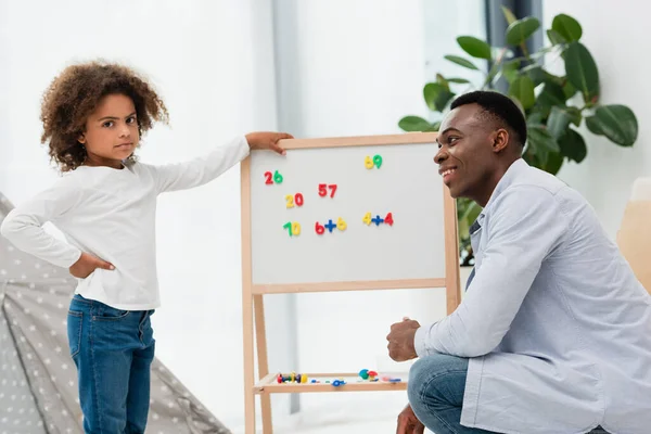 Afrikanisch-amerikanischer Vater schaut ernste Tochter an, die mit der Hand auf Hüfte neben Whiteboard mit Magneten steht — Stockfoto