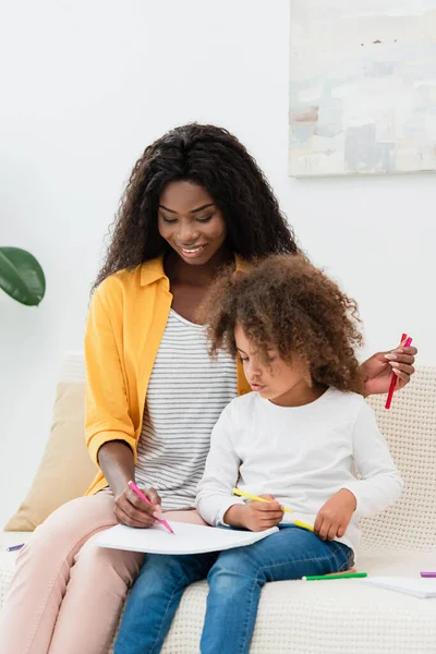 Afro-américaine mère dessin avec crayon de couleur tout en étant assis sur le canapé avec fille bouclée — Photo de stock