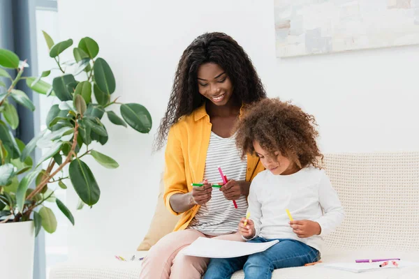 Afro-américaine mère tenant crayon de couleur tout en étant assis sur le canapé avec fille bouclée — Photo de stock