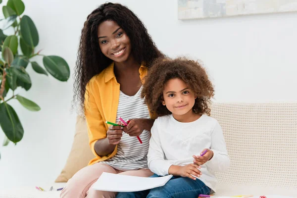 Madre afroamericana y su hija sosteniendo lápiz de color mientras están sentadas en el sofá - foto de stock