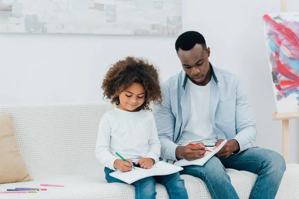 African american father and daughter drawing with color pencils while sitting on sofa — Stock Photo