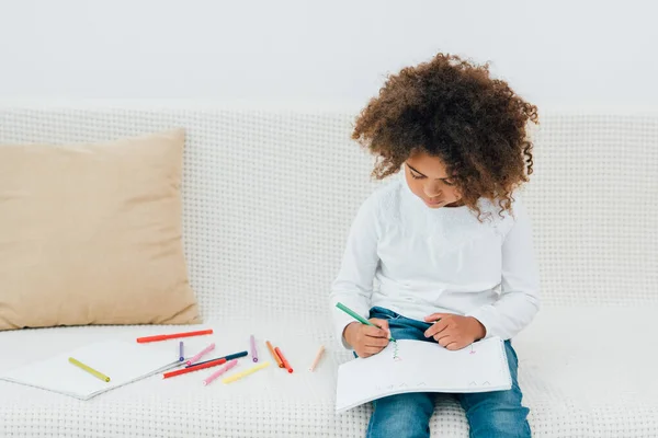 Niño afroamericano rizado sentado en el sofá y dibujo con lápiz de color - foto de stock