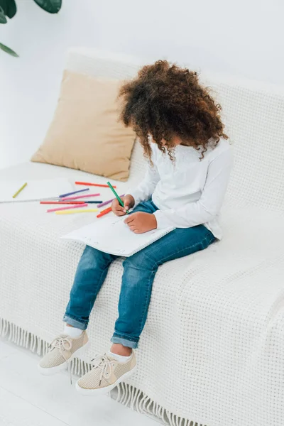 Curly african american kid drawing with color pencil while sitting on sofa — Stock Photo