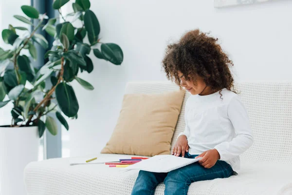 Ricci bambino africano americano guardando carta bianca mentre seduto sul divano vicino alle matite di colore — Foto stock