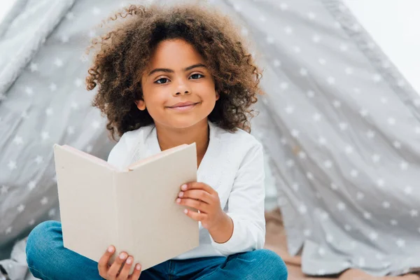 Lockiges afrikanisch-amerikanisches Kind hält Buch in der Hand und blickt in die Kamera — Stockfoto