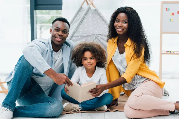 African american parents and curly kid holding book while sitting on floor — Stock Photo