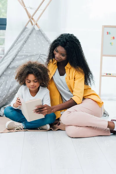 African american mother and daughter sitting on floor and reading book — Stock Photo