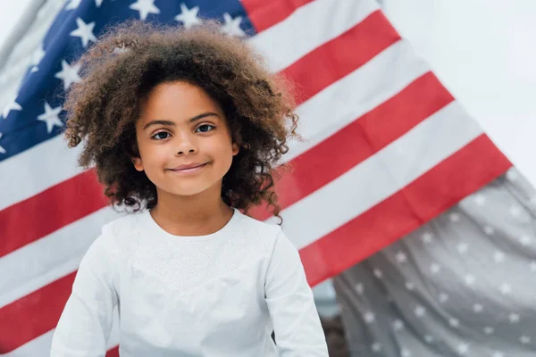 Curly african american kid looking at camera near flag of america — Stock Photo
