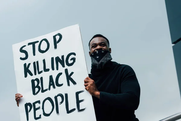 Homme afro-américain avec écharpe sur le visage tenant la pancarte avec cesser de tuer les personnes noires lettrage à l'extérieur — Photo de stock