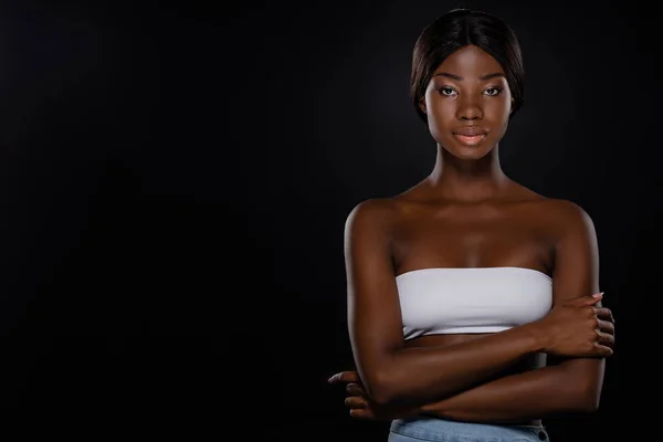 Mujer afroamericana con brazos cruzados mirando a la cámara aislada en negro - foto de stock