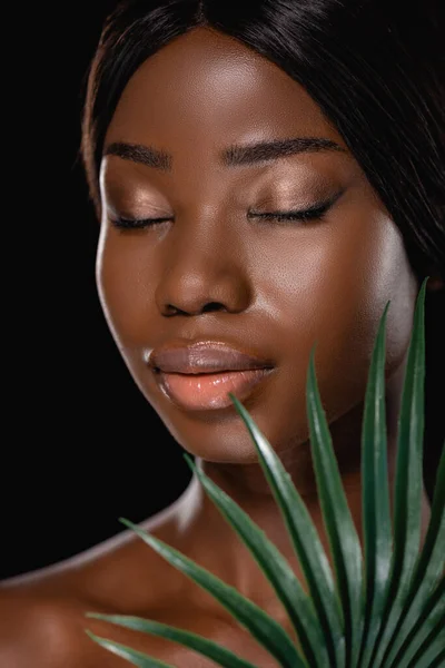 Mujer desnuda afroamericana con los ojos cerrados y hoja de palma verde aislado en negro — Stock Photo