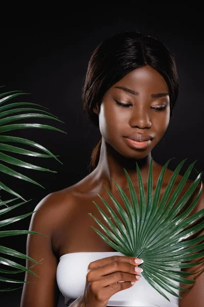 African american woman near green palm leaves isolated on black — Stock Photo