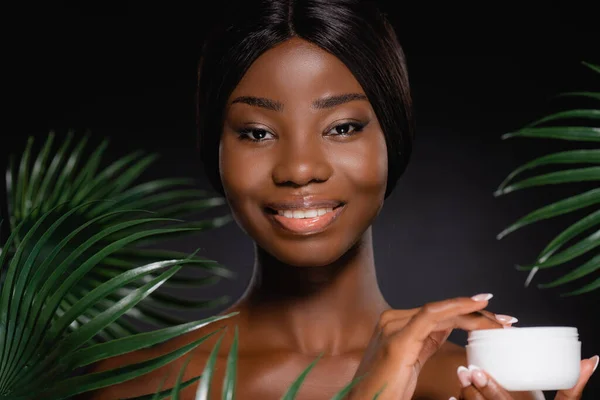 African american naked woman with cosmetic cream near green palm leaves isolated on black — Stock Photo