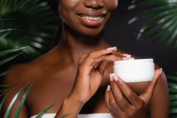 Vista ritagliata di donna afro-americana applicando crema cosmetica vicino foglie di palma verde isolato su nero — Foto stock