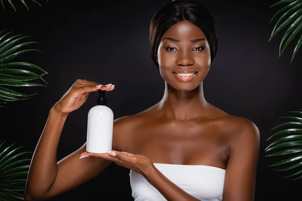 African american woman holding body lotion in dispenser bottle near green palm leaves isolated on black — Stock Photo