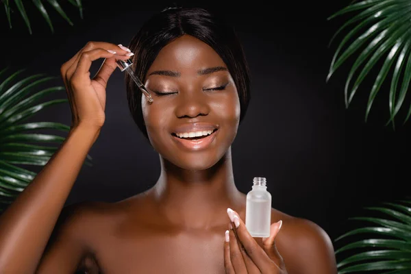 African american naked woman applying serum on face near green palm leaves isolated on black — Stock Photo