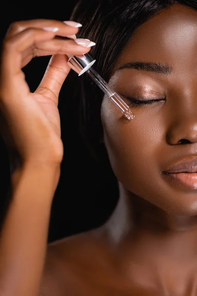 Porträt einer nackten afrikanisch-amerikanischen Frau, die Serum auf das Gesicht aufträgt, isoliert auf schwarz — Stockfoto