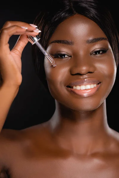 Retrato de mujer desnuda afroamericana aplicando suero en la cara aislada en negro - foto de stock