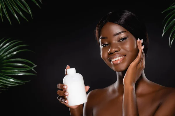 Africano americano mulher nua segurando garrafa de loção perto de folhas de palma verde isolado em preto — Fotografia de Stock