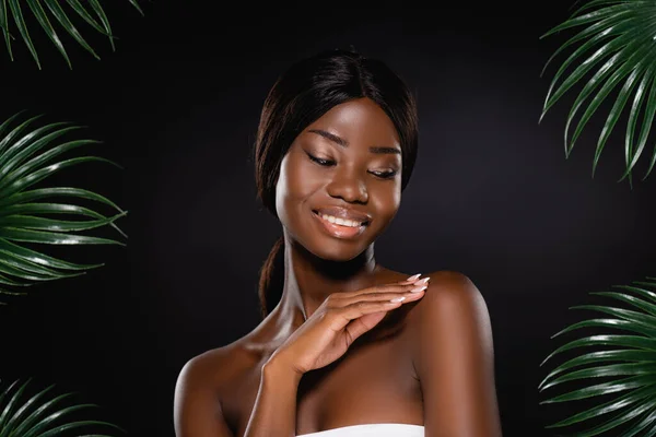 Mujer afroamericana tocando cuerpo cerca de hojas de palma verde aisladas en negro - foto de stock