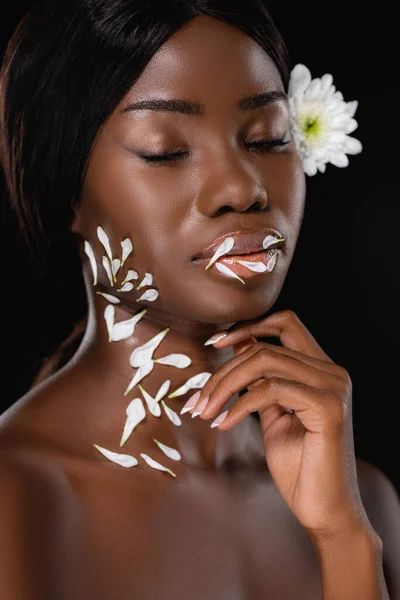 African american woman with closed eyes, white chrysanthemum in hair and petals on neck and lips isolated on black — Stock Photo