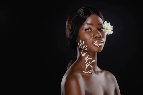 Naked african american woman with white chrysanthemum in hair and petals on neck and lips isolated on black — Stock Photo