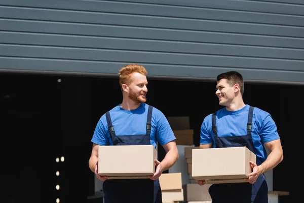 Young movers in uniform looking at each other while holding cardboard boxes on urban street — Stock Photo