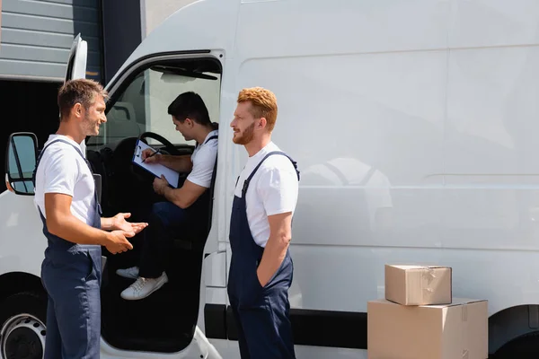 Movers in uniform talking near colleague writing on clipboard in trunk — Stock Photo