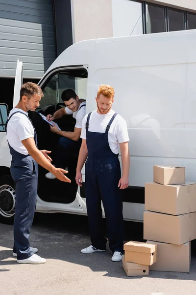Movers in overalls looking at cardboard boxes near colleague in truck on urban street — Stock Photo