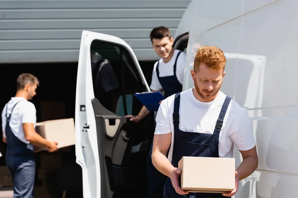 Selective focus of loader holding cardboard box while colleagues working near truck on urban street — Stock Photo