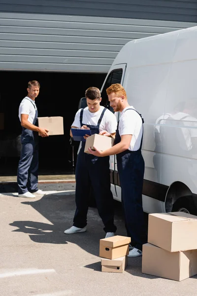 Selective focus of mover holding cardboard box near colleague writing on clipboard and truck on urban street — Stock Photo