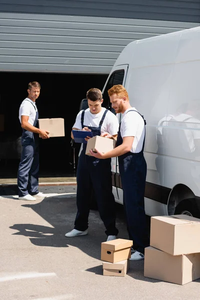 Focus selettivo del traslocatore con scatola di cartone vicino al collega che scrive sugli appunti e sul camion sulla strada urbana — Foto stock