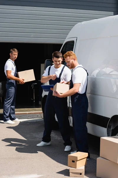 Selective focus of mover pointing at cardboard box near colleague with pen and clipboard beside truck on urban street — Stock Photo