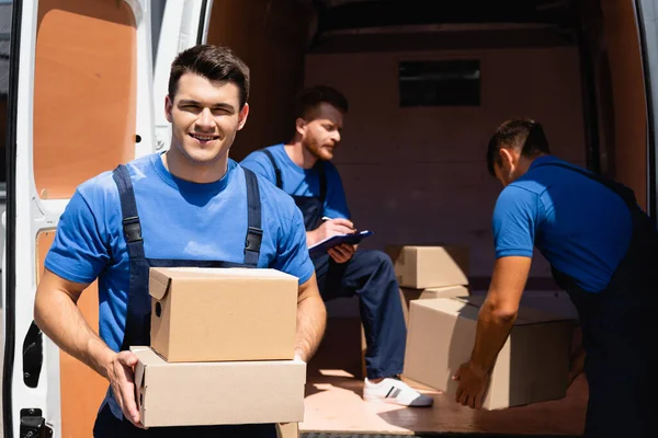 Selective focus of young loader holding cardboard boxes and looking at camera while colleagues unloading truck outdoors — Stock Photo