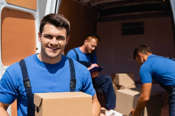 Selektiver Fokus des Laders mit Pappkartons, der in die Kamera schaut, während Kollegen in der Nähe des Lastwagens im Freien arbeiten — Stockfoto