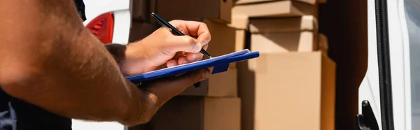 Cultivo panorámico de la escritura del cargador en portapapeles con cajas de cartón en el camión en el fondo - foto de stock