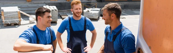 Panoramic shot of loaders looking at each other near door of truck on urban street — Stock Photo