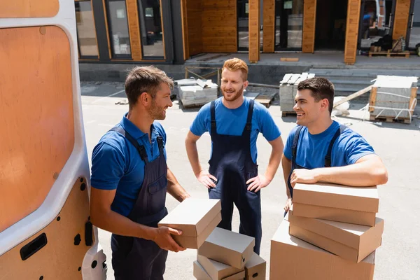 Movers in overalls looking at each other near carton boxes and truck on urban street — Stock Photo