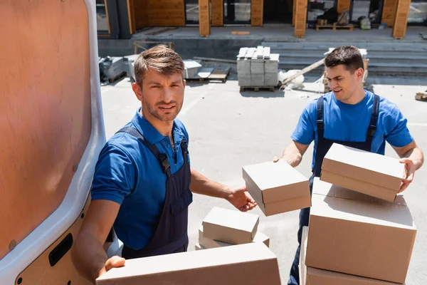 Selective focus of loader looking at camera while holding carton box near colleague and truck on urban street — Stock Photo