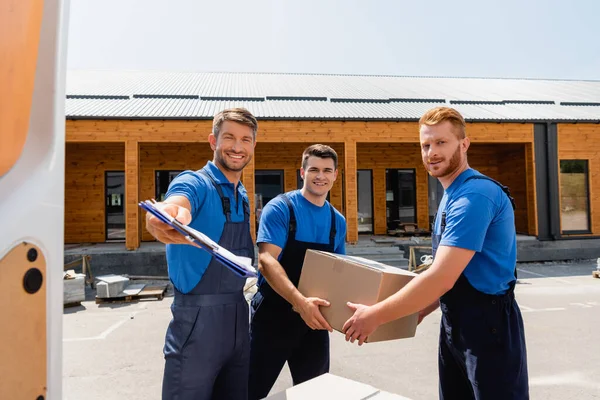 Selective focus of loader holding clipboard near colleagues with cardboard box and door of truck on urban street — Stock Photo