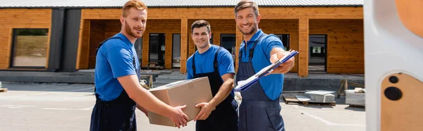 Horizontal crop of movers with clipboard and cardboard box looking at camera near door of truck on urban street — Stock Photo