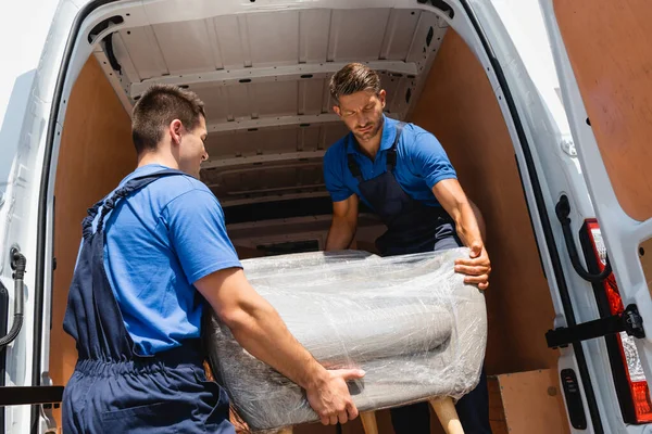 Movers in uniform loading armchair in stretch wrap in truck outdoors — Stock Photo