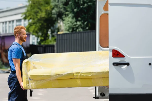 Side view of loader in uniform holding couch in stretch wrap near truck with open doors on urban street — Stock Photo