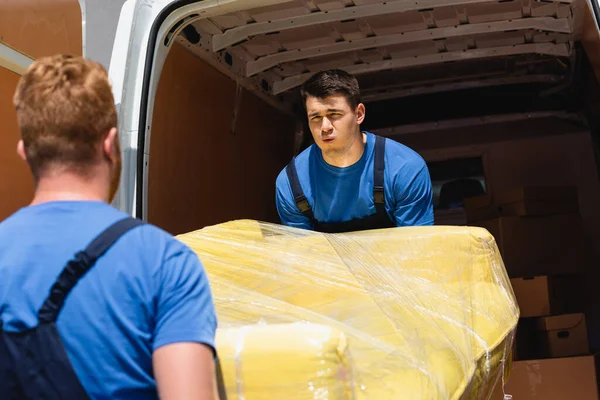 Selective focus of tired loader carrying couch in stretch wrap with coworker near truck with open doors outdoors — Stock Photo