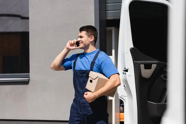 Selective focus of loader holding carton box and talking on smartphone near truck on urban street — Stock Photo