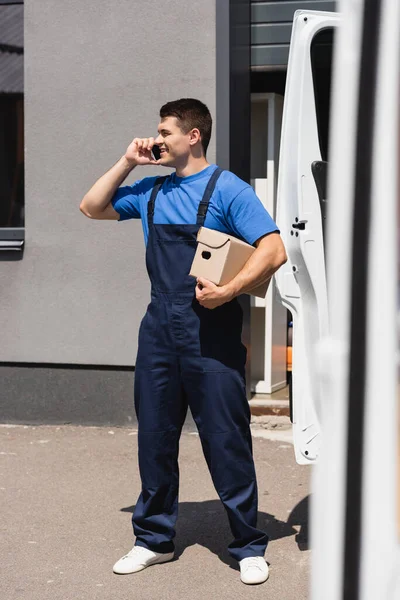 Selective focus of loader in overalls talking on smartphone and holding package near truck on urban street — Stock Photo